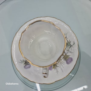 Könnte beinhalten: Eine weiße Teetasse und Untertasse mit goldfarbenem Rand und floralem Design. Die Untertasse zeigt eine violette Distelblüte und grüne Blätter.