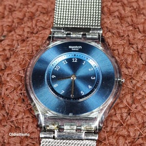 May include: A Swatch Swiss watch with a blue dial and silver-tone mesh band. The watch face features a radial pattern and white numerals. The watch has a clear plastic case and a silver-tone metal band. The watch is a vintage item.