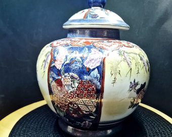 Puede incluir: Un jarrón de cerámica decorativo con tapa, con un detallado patrón floral y geométrico en rojo, azul y dorado. El jarrón tiene forma redonda con una base plateada y un botón azul en la tapa.