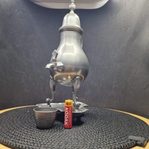 以下が含まれることがあります： 蓋、注ぎ口、3本足のヴィンテージピューターティーポットまたはコーヒーポット。小さなピューターカップとKodak AAバッテリーがポットの前にあります。ポットは装飾的なトレイの上にあります。