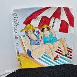 May include: A ceramic serving platter with a painted scene of two women in swimsuits relaxing on beach chairs under a red and white striped umbrella. The text "Girlfriends in summertime" is written on the side of the platter.