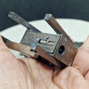 May include: A vintage, bronze-colored pencil sharpener with a rectangular body and hinged arms. The sharpener has the text "MADE IN" and "N-1004" on its surface. The design includes a circular hole and a small screw.