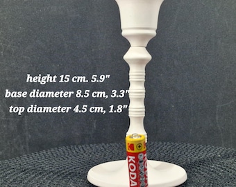 May include: A white candlestick holder with a round base and a decorative stem. The top diameter is 4.5 cm, the base diameter is 8.5 cm, and the height is 15 cm. A red and yellow Kodak battery is placed next to the holder.
