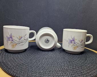 May include: Three white ceramic mugs with a floral design featuring purple and yellow flowers. The mugs have a simple, classic shape with a handle on the side. The mugs are arranged on a dark grey surface.