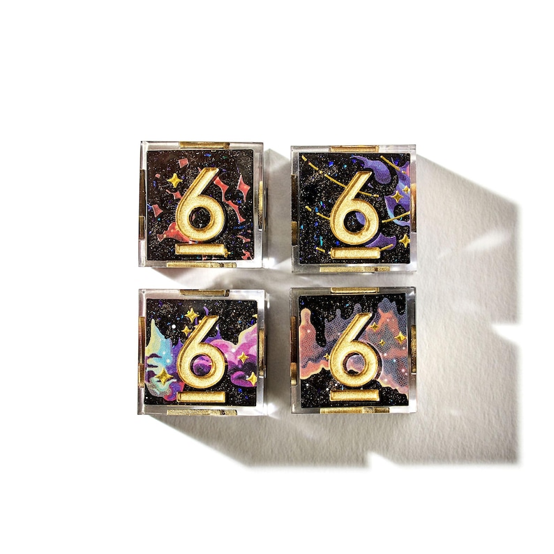 May include: Four square compacts with a gold number 6 on a black background with colorful accents. The compacts are in clear cases with gold trim. The background has a galaxy theme with stars and colorful nebula-like designs.