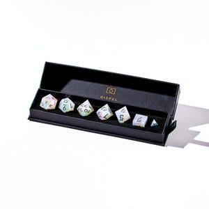 May include: A set of seven iridescent dice in a black box with the word "DISPEL" printed on the box. The dice are in a variety of shapes and sizes, and they are all made of a clear material with a rainbow sheen.