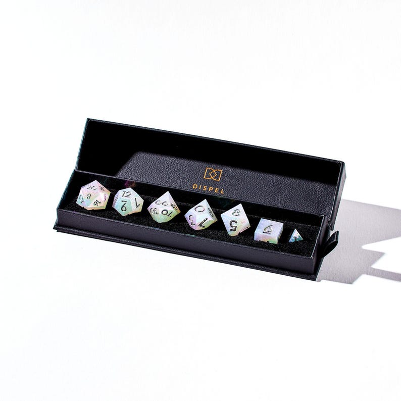 May include: A set of seven iridescent dice in a black box with the word "DISPEL" printed on the box. The dice are in a variety of shapes and sizes, and they are all made of a clear material with a rainbow sheen.