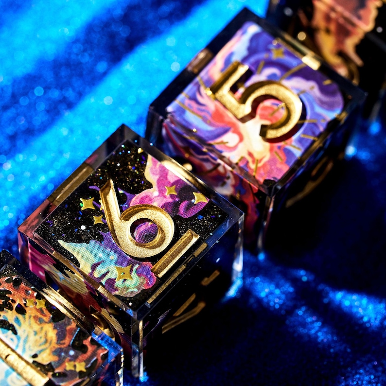 May include: Close-up of several dice with a celestial theme. Each die features a gold number, such as 6 and 5, set against a colorful, swirling background of purples, pinks, and blues. The dice are encased in clear resin with gold accents.