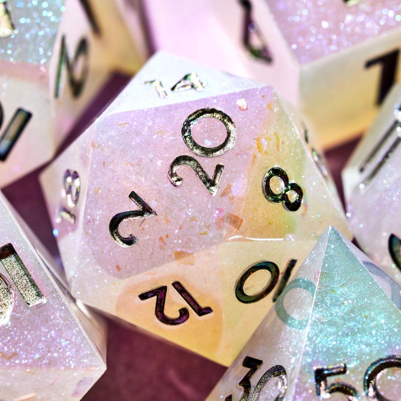 May include: Close-up of a pink and white iridescent 20-sided die with gold numbers. The die is made of resin and has a glittery finish.