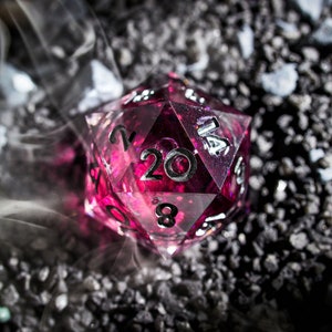 以下が含まれることがあります： ピンクと透明な樹脂ボディのD20サイコロのクローズアップ。サイコロにはシルバーの数字があり、濃い灰色の砂利の上に置かれています。背景には白い煙が漂っています。