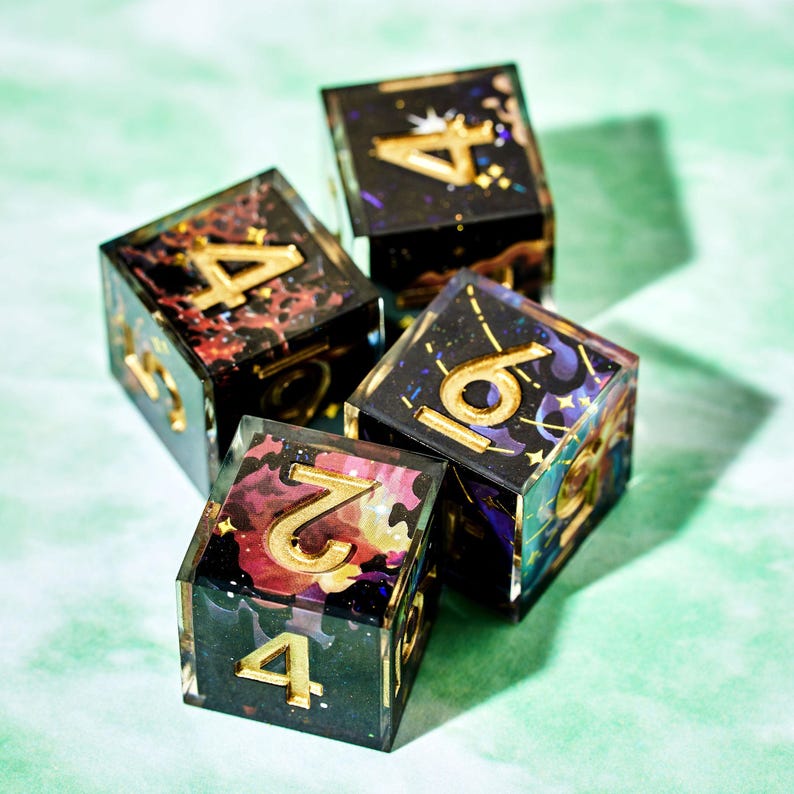 May include: Four square dice with gold numbers and colorful, galaxy-like designs inside. The dice are black with gold numerals, and the interior features swirls of red, purple, and gold. The dice are arranged on a light green surface.