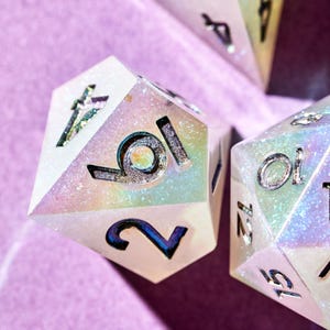 May include: Close-up of a white iridescent dice with silver numbers. The dice shows the numbers 10 and 2. The dice is on a pink surface.