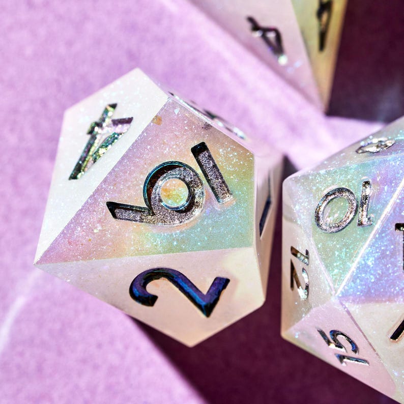 May include: Close-up of a white iridescent dice with silver numbers. The dice shows the numbers 10 and 2. The dice is on a pink surface.