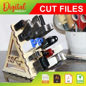 以下が含まれることがあります： 三角形の形状をした木製スタンドで、前面にUSBのシンボルが彫刻されています。スタンドはUSBドライブを収納するために設計されており、スタンドにはさまざまな色とサイズのUSBドライブがいくつかあります。スタンドは黒い表面に置かれており、背景には白いボウル、ハサミ、虫眼鏡などがあります。画像には「CUT FILES」と「SVG、AI、PDF、CDR」というテキストが含まれています。