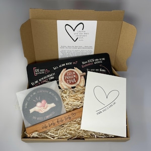 Könnte beinhalten: Eine Geschenkbox mit verschiedenen Artikeln, darunter ein Untersetzer, ein Holzlesezeichen, eine Grußkarte und eine dekorative Tafel. Die Artikel haben Herzmotive und deutschen Text. Die Box ist mit Stroh gefüllt.