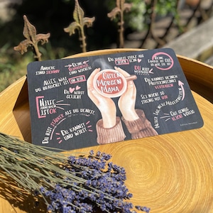 Könnte beinhalten: Ein schwarzes rechteckiges Schild mit deutschem Text, darunter "Guten Morgen Mama". Das Schild zeigt eine Aquarellillustration von Händen, die eine Kaffeetasse halten. Das Schild liegt auf einem Holztablett mit Lavendel.