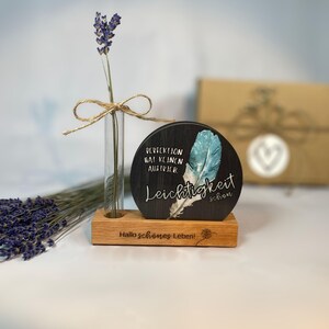 Könnte beinhalten: Ein Holzdisplay mit einer kleinen Glasvase mit einem Lavendelzweig, der mit Bindfaden gebunden ist, und einer runden schwarzen Tafel mit einem blauen Federdesign und deutschem Text. Die Basis ist mit Text graviert.