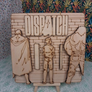 May include: Wooden laser-cut artwork featuring the word "DISPATCH" above three figures. The figures are standing in front of a brick wall design. The artwork is displayed on a small wooden easel.