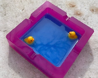 May include: A pink resin ashtray shaped like a pool with two yellow rubber duckies floating in the blue water.