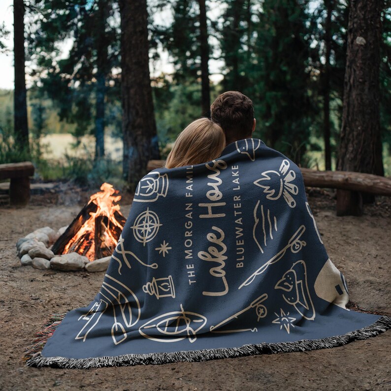 May include: A navy blue throw blanket with a cream-colored fringe. The blanket features a design with camping-themed illustrations and the text "Lake House Bluewater Lake". The blanket is draped over two people sitting near a campfire.