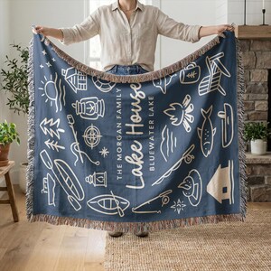 May include: A navy blue throw blanket with a cream-colored fringe. The blanket features white line art illustrations of lake-themed icons and the words "Lake House" and "Bluewater Lake". The blanket is rectangular.