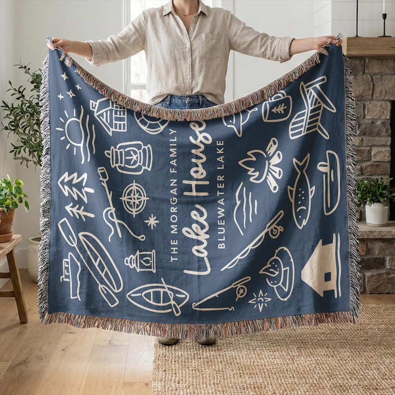 May include: A navy blue throw blanket with a cream-colored fringe. The blanket features white line art illustrations of lake-themed icons and the words "Lake House" and "Bluewater Lake". The blanket is rectangular.