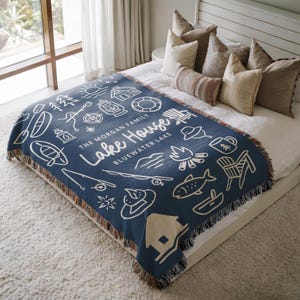 May include: A navy blue woven throw blanket with a white illustrated lake house design. The blanket features various outdoor icons, including a sun, trees, and a campfire. Text on the blanket reads "Lake House" and "Bluewater Lake".