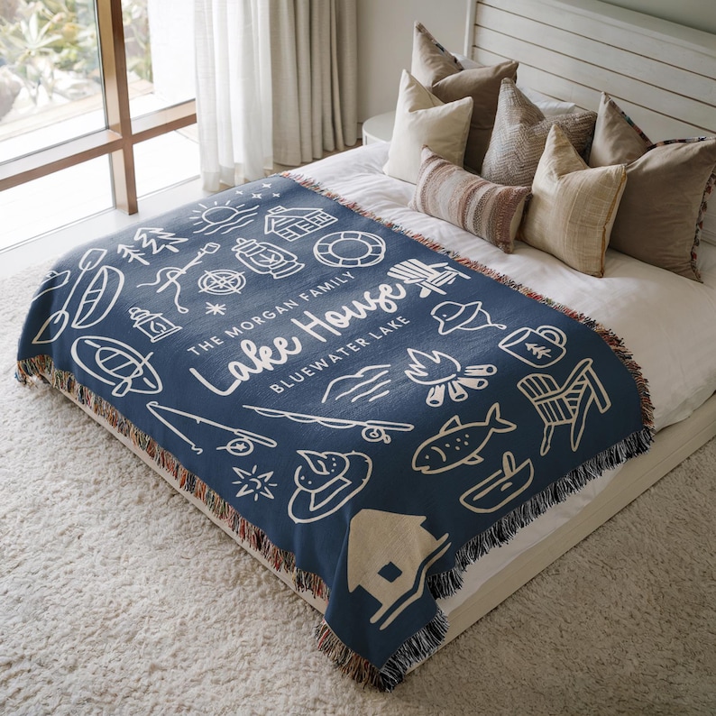 May include: A navy blue woven throw blanket with a white illustrated lake house design. The blanket features various outdoor icons, including a sun, trees, and a campfire. Text on the blanket reads "Lake House" and "Bluewater Lake".