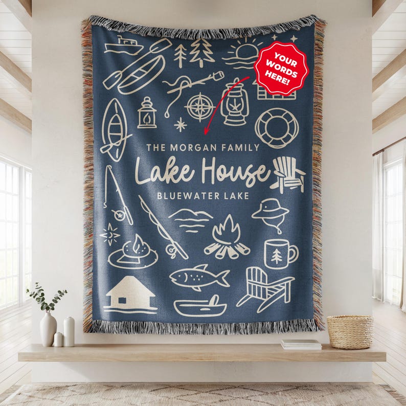 May include: A navy blue woven throw blanket with a cream-colored lakeside design. The blanket features illustrations of a boat, fishing rod, campfire, and the text "Lake House, Bluewater Lake". The blanket has a fringed edge.