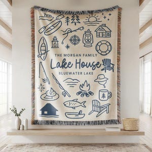 May include: Cream-colored woven throw blanket with a navy blue lake house design. The design includes illustrations of a boat, fishing rod, campfire, and the words "Lake House" and "Bluewater Lake". The blanket has a fringed edge.