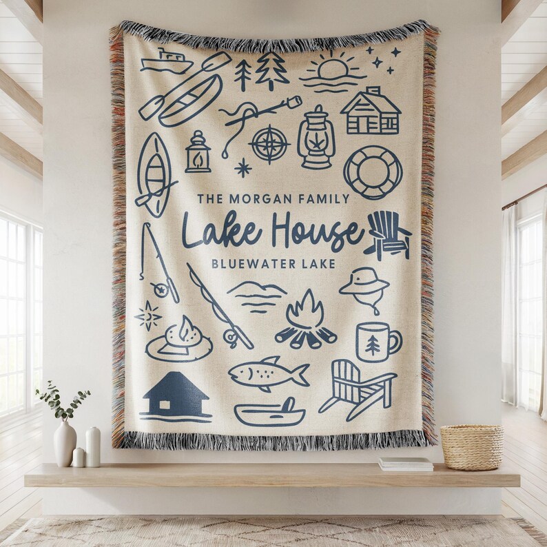May include: Cream-colored woven throw blanket with a navy blue lake house design. The design includes illustrations of a boat, fishing rod, campfire, and the words "Lake House" and "Bluewater Lake". The blanket has a fringed edge.