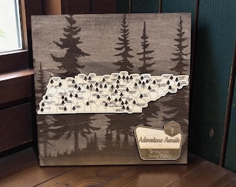 Puede incluir: Arte de pared de madera con un mapa de Tennessee con pequeños marcadores de árboles. El fondo tiene un diseño de silueta de bosque. Un pequeño letrero dice "Adventure Awaits Tennessee State Parks."