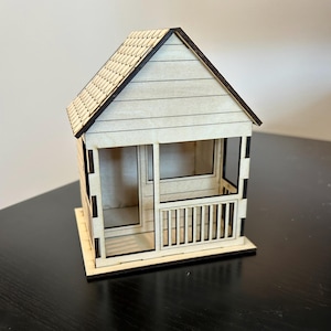 May include: A small wooden model of a house with a porch and a shingled roof. The house is made of light wood and has a dark brown trim.