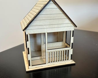 May include: A small wooden model of a house with a porch and a shingled roof. The house is made of light wood and has a dark brown trim.