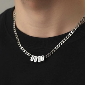 Puede incluir: Un collar de cadena de plata con cuatro cuentas rectangulares blancas. Cada cuenta tiene un nombre impreso en letras negras: Mary, Jack, Rose y Dan. El collar se usa sobre una camiseta negra.