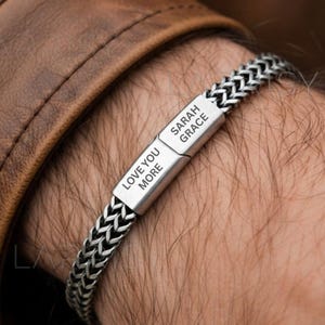 Puede incluir: Una pulsera plateada con un diseño de cadena trenzada. La pulsera presenta un cierre rectangular grabado con las palabras "LOVE YOU MORE" y "SARAH GRACE". La pulsera se lleva en la muñeca, con una chaqueta de cuero marrón en el fondo.