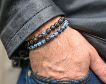 May include: Two beaded bracelets, one black and one blue, are worn on a wrist. The black bracelet features a textured, porous design, while the blue bracelet has a speckled pattern. The bracelets are displayed against a background of a black leather jacket and blue jeans.