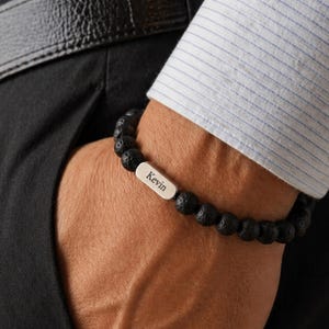 Peut inclure: Bracelet de perles de lave noires avec une plaque nominative argentée gravée du nom "Kevin". Le bracelet est porté au poignet, la personne portant une chemise rayée blanche et bleue et un pantalon noir. Une ceinture noire est également visible.