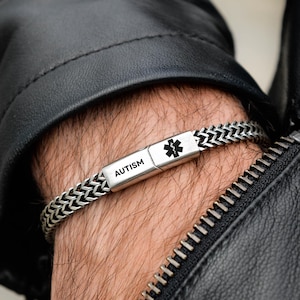May include: A silver-toned medical alert bracelet featuring a woven chain design and a rectangular metal plate. The plate is engraved with the word AUTISM and a black Star of Life medical symbol. The bracelet has a secure clasp mechanism.