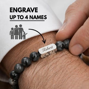 May include: A beaded bracelet with a silver rectangular charm engraved with the name "Robert" and two heart symbols. The bracelet is made of dark grey, round beads. Text above the bracelet reads "ENGRAVE UP TO 4 NAMES".