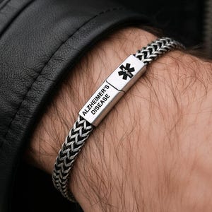 Puede incluir: Una pulsera de identificación médica plateada con un diseño de cadena trenzada. La pieza central rectangular está grabada con "ALZHEIMER'S DISEASE" y un símbolo médico. La pulsera se lleva en la muñeca.