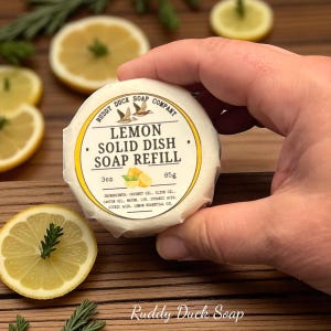 以下が含まれることがあります： 「Ruddy Duck Soap Company」の文字が入った、手で持たれた丸く白いレモンソリッド食器用石鹸詰め替え用。ラベルには「Lemon Solid Dish Soap Refill」と書かれ、成分が記載されています。詰め替え用は85g（3オンス）です。