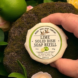 以下が含まれることがあります： 「Ruddy Duck Soap」と「Lime Solid Dish Soap Refill」の文字が入った、手に持たれた丸いライムの香りの固形食器用石鹸詰め替え用。ラベルにはライムのグラフィックと成分が記載されています。ライムと花が背景にあります。