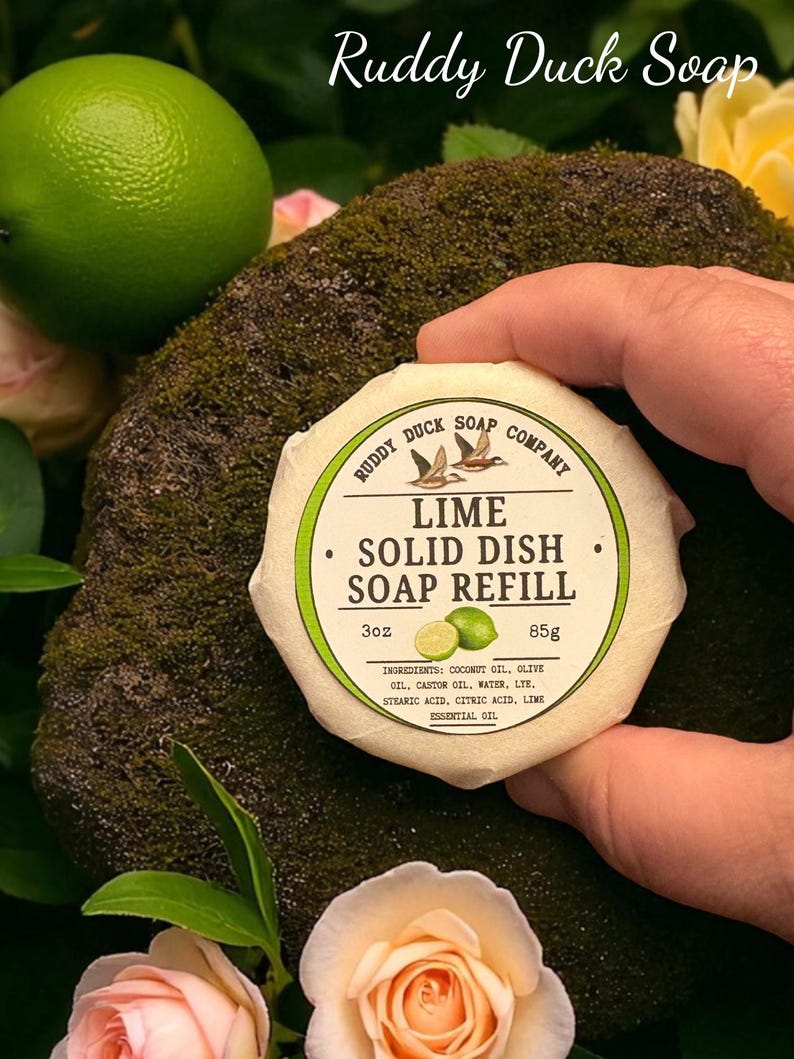 以下が含まれることがあります： 「Ruddy Duck Soap」と「Lime Solid Dish Soap Refill」の文字が入った、手に持たれた丸いライムの香りの固形食器用石鹸詰め替え用。ラベルにはライムのグラフィックと成分が記載されています。ライムと花が背景にあります。