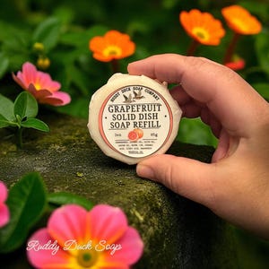 May include: A round grapefruit solid dish soap refill from Ruddy Duck Soap Company. The label features the text "Grapefruit Solid Dish Soap Refill" and a graphic of a bird. The soap weighs 3 oz and is held in front of a background of pink and orange flowers.