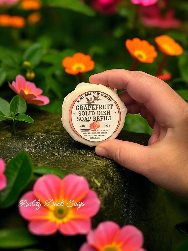 以下が含まれることがあります： Ruddy Duck Soap Companyのグレープフルーツ固形食器用石鹸詰め替え用。ラベルには「Grapefruit Solid Dish Soap Refill」の文字と鳥のグラフィックが描かれています。石鹸の重量は85gで、ピンクとオレンジの花を背景に持っています。