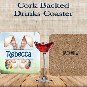 May include: A square cork-backed drinks coaster with an Easter theme. The coaster has a design with bunny ears, the name Rebecca, and Easter eggs. A wine glass with red wine is next to the coaster. The back of the coaster is cork.