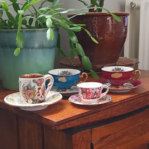 Puede incluir: Cuatro tazas de té de porcelana con intrincados diseños florales y dorados. Las tazas están dispuestas sobre una mesa de madera con un cajón.