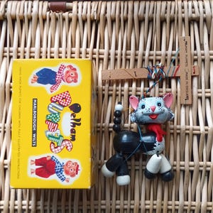 May include: A vintage Pelham puppet cat with a red scarf, black body, and white feet, next to a yellow box with the text "Selham" and illustrations. The puppet is controlled by strings attached to a wooden cross.
