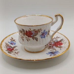 Puede incluir: Un juego de taza y platillo de té blanco con un patrón floral en tonos rojos, azules y verdes. El juego tiene un borde dorado.